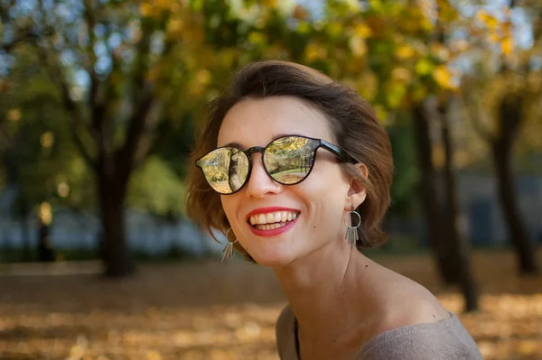 Ung och vacker flicka med kort hår och solglasögon med spegelyta poserar mot träden med gula blad bakgrund tillbringa tid i höstparken — Stockfoto