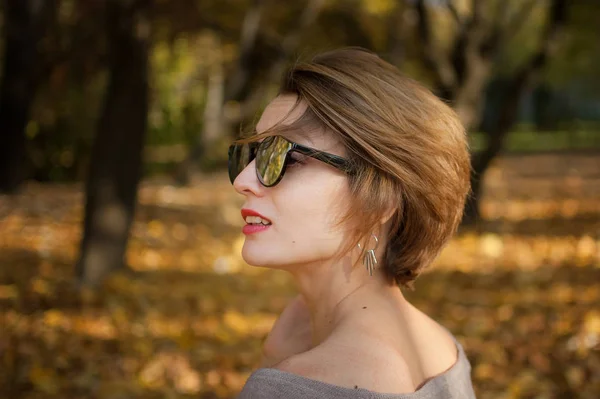 Menina jovem e bonita com cabelo curto e óculos de sol com superfície de espelho está posando contra as árvores com folhas amarelas fundo passar o tempo no parque de outono — Fotografia de Stock