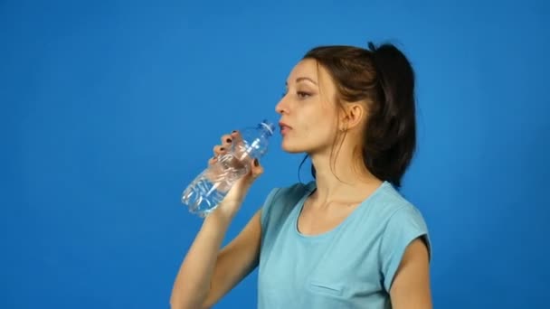 Vue latérale de la fille brune aux cheveux longs et foncés boit une eau pure à partir d'une bouteille transparente en plastique après son entraînement sur un fond bleu en studio — Video