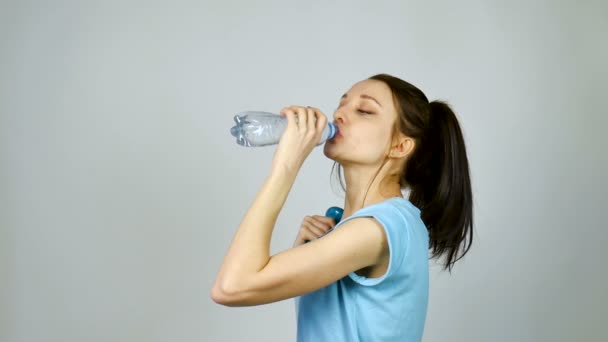 Brunette κορίτσι με μακριά σκούρα μαλλιά πίνει καθαρό νερό από πλαστικό διαφανές μπουκάλι μετά την προπόνηση της σε ένα γκρι φόντο στο στούντιο — Αρχείο Βίντεο