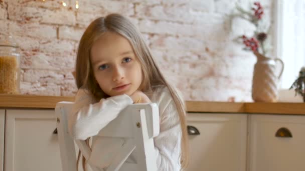 Petite fille triste aux yeux bleus et aux longs cheveux blonds portant une robe blanche et un pull s'assoit sur une chaise dans une pièce de style loft — Video