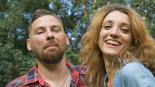 Preciosa pareja de novio hipster barbudo en camisa roja y novia rubia con pantalones vaqueros azules casuales mirando a la cámara en el fondo de los árboles verdes — Vídeos de Stock