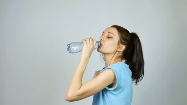 Brunetka dziewczyna z długimi ciemnymi włosami pije czystą wodę z plastikowej przezroczystej butelki po treningu na szarym tle w studio — Wideo stockowe