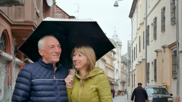 Middle-aged woman and her elderly husband spending time together outdoors standing under their umbrella on paved street. Couple with age difference. — Stock Video