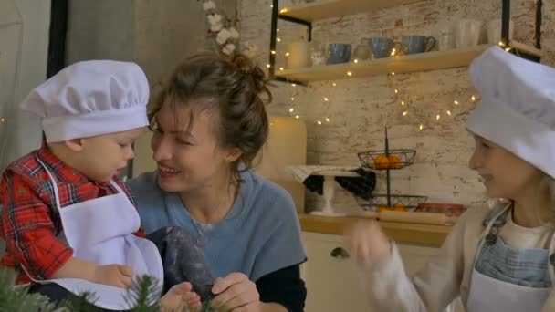 Νεαρή σγουρή μητέρα με το γιο και την κόρη της μαγειρεύουν χριστουγεννιάτικα μπισκότα στο σπίτι στην κουζίνα με χριστουγεννιάτικη διακόσμηση. — Αρχείο Βίντεο