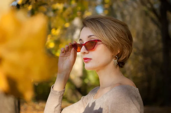 Menina jovem e bonita com cabelo curto e óculos de sol na moda vermelho está posando contra as árvores com folhas amarelas fundo passar o tempo no parque de outono — Fotografia de Stock