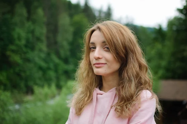 Atractiva joven mujer rubia tranquila en ropa rosa con pelo largo en la colina con fondo de bosque verde durante la primavera o principios de otoño en las montañas . — Foto de Stock