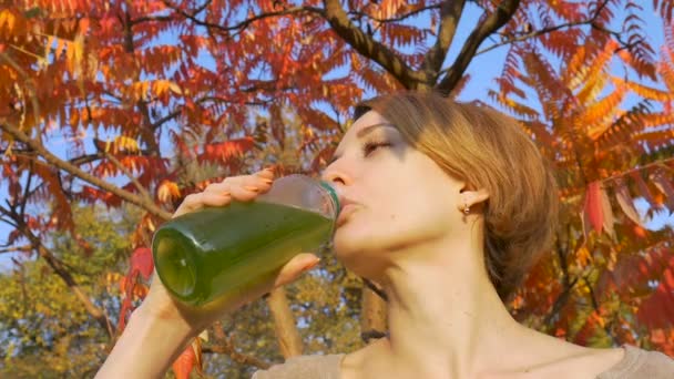 La mujer está bebiendo bebida verde, espirulina, clorella y batido de hierba de trigo al aire libre durante el soleado día de otoño sobre un fondo de árbol con hojas rojas y naranjas. Estilo de vida saludable, desintoxicación . — Vídeo de stock