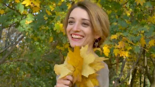 Emocional joven atractiva con cabello corto rubio y vestido biege sosteniendo un ramo de hojas de otoño por encima de su cabeza y posando en un hermoso parque al aire libre — Vídeos de Stock