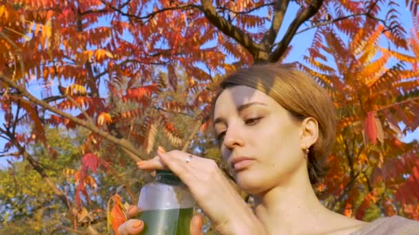 Hermosa chica está bebiendo bebida verde, espirulina, clorella y batido de hierba de trigo al aire libre en el parque sobre un fondo de árbol con hojas rojas y naranjas. Estilo de vida saludable, desintoxicación . — Vídeo de stock