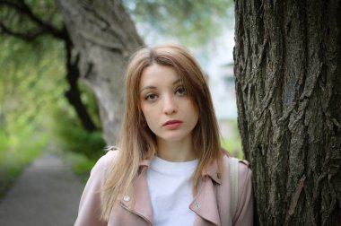 Açık havada, bir bahar parkında büyük bir ağacın yanında poz veren sarı saçlı çekici bir kadının kadın portresi. Ağaca yaslanır ve kabuğuna dokunur.
