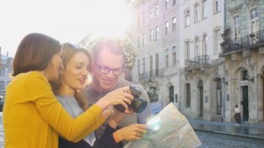 Üç mutlu genç turist, sabahın erken saatlerinde boş bir meydanda antik Avrupa şehrinin kağıt haritasını tutuyor ve fotoğraflarına bakıyorlardı.