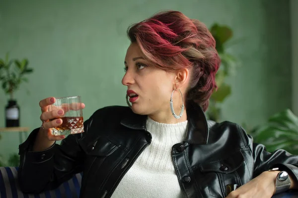 Junge traurige Frau mit dunkelrosa Haaren mit Glam-Rock-Look trinkt Whisky aus einem Glas mit abscheulicher Fratze im Gesicht, weiblicher Alkoholismus, Alkoholsucht — Stockfoto