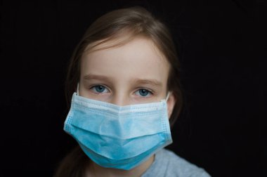 Coronavirus Covid-19 salgını. Stüdyoda siyah arka planda virüsü korumak için açık mavi tek kullanımlık maske takan küçük sarışın kız. Karantina konsepti