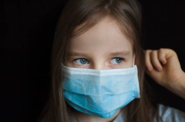 Kendini izole ederken Coronavirus Covid-19 'un korunması için mavi tek kullanımlık maskeli siyah arka plandaki küçük sarışın kızın stüdyo portresi.