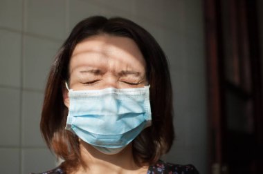 Kendini tecrit sırasında Coronavirus Covid-19 'dan korunmak için yüzünde tek kullanımlık maske olan bir kadın portresi ve mutfakta arka planda beyaz fayansların üzerinde kapalı gözlerle kalmak.