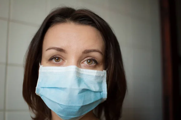 Sad adult girl is restricted at home because of the pandemia of Coronavirus Covid-19 and self-isolation. Woman is at the kitchen during sunny day on white tiles background in disposable mask — Stock Photo, Image