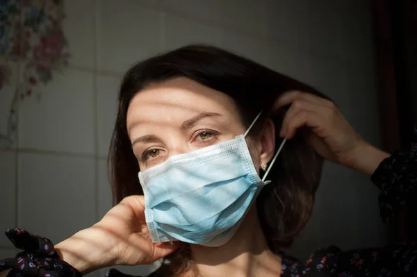 Woman is trying put the disposable mask on her face to protect herself from Coronavirus Covid-19 during self-isolation and stay home on white tiles background at the kitchen — Stock Photo, Image