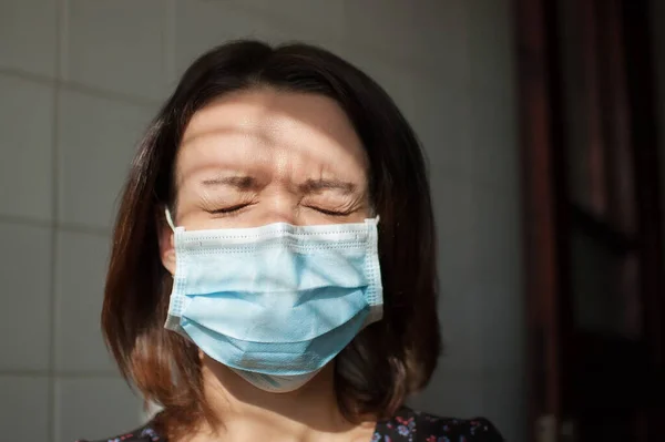 Retrato femenino de mujer con máscara desechable en la cara para protegerse del Coronavirus Covid-19 durante el autoaislamiento y quedarse en casa sobre fondo de baldosas blancas en la cocina con los ojos cerrados —  Fotos de Stock