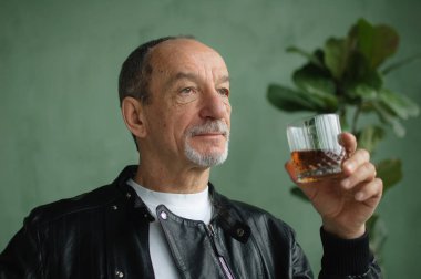 Son sınıf öğrencisinin elinde viski ya da brendi bardağı tutarken ve arka planda açık yeşil duvarlar ve ev bitkileri olan tavan arası tarzı bir odada bir şeyler düşünürken yan görüntüsü. Alkol bağımlılığı