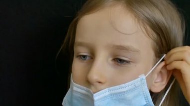 Mavi gözlü ve uzun saçlı küçük sarışın kız evde yalnızken Coronavirus Covid-19 'un korunması için ameliyat maskesiyle siyah arka planda poz veriyor.