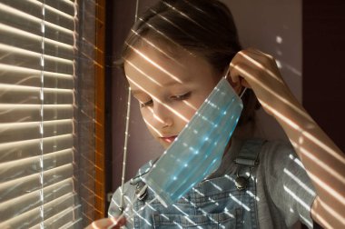Küçük kız pencere eşiğinde oturuyor ve perdelerin arkasındaki Coronavirus Covid-19 yüzünden kendini izole ederken yüzüne tek kullanımlık koruyucu maske takmaya çalışıyor.