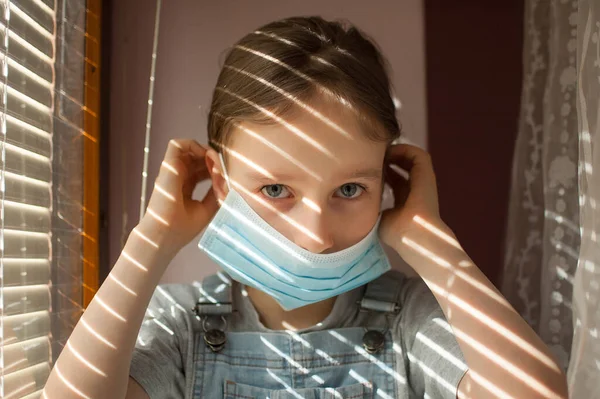 Niña está sentada en el alféizar de la ventana y tratando de poner máscara protectora desechable en su cara durante el auto-aislamiento en casa debido a Coronavirus Covid-19 en el fondo de persianas — Foto de Stock