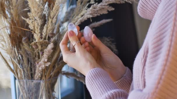 Mãos femininas segurando dois ovos yoni feitos de quartzo rosa e ametista violeta transparente para vumfit, imbuilding ou meditação perto da janela com vaso de espiguetas . — Vídeo de Stock