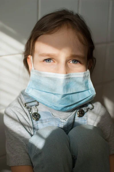 Little sad girl is restricted at home because of the pandemia of Coronavirus Covid-19 and self-isolation. Kid is sitting at the kitchen on white tiles background in a protective disposable mask — Stock Photo, Image