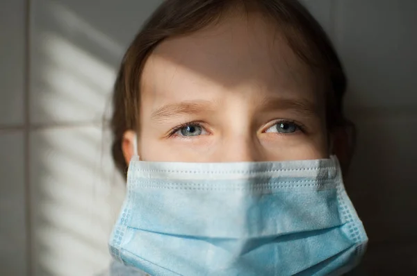 Retrato de la niña triste en una máscara protectora desechable se queda en casa debido a la pandemia de Coronavirus Covid-19 en auto-aislamiento sobre fondo de baldosas blancas en la cocina durante el día soleado — Foto de Stock