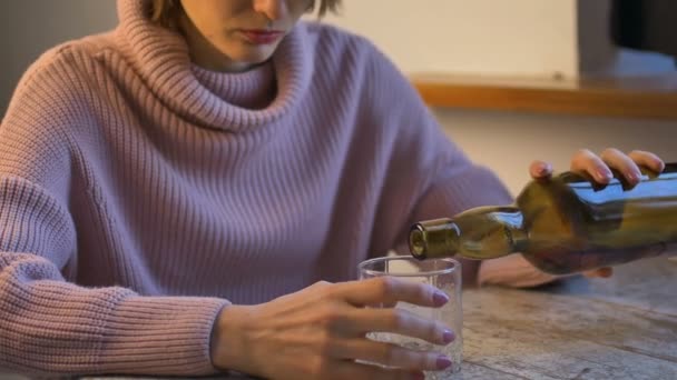 Mujer triste en jeans y suéter rosa está sentado en la mesa y beber un whisky del vaso, botella verde oscuro es visible, alcoholismo femenino — Vídeos de Stock