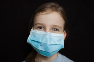 Coronavirus Covid-19 salgını. Stüdyodaki siyah arka planda virüsü korumak için mavi gözlü küçük sarışın kız tek kullanımlık maske takıyor. Kişisel koruyucu ekipman konsepti