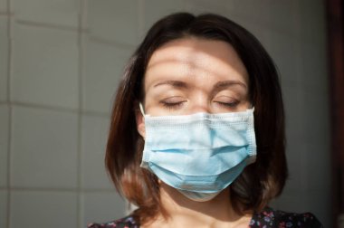 Kendini tecrit sırasında Coronavirus Covid-19 'dan korunmak için yüzünde tek kullanımlık maske olan bir kadın portresi ve mutfakta arka planda beyaz fayansların üzerinde kapalı gözlerle kalmak.