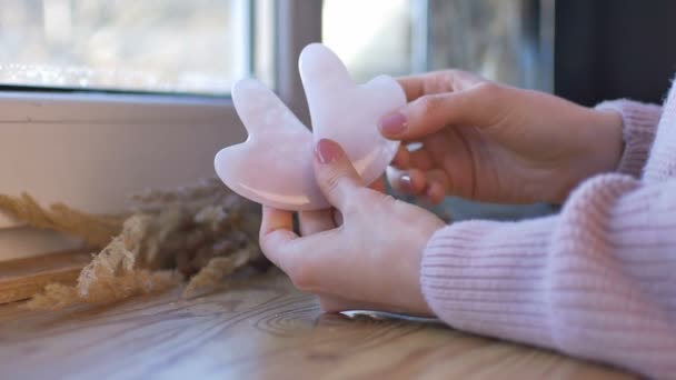 Las manos femeninas con manicura sostienen la herramienta de masaje gua sha hecha de piedra natural de cuarzo rosa cerca de la ventana. Elevación, cuidado natural y desintoxicación — Vídeo de stock