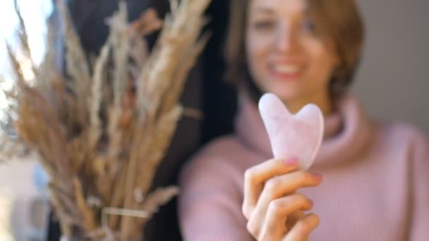 A menina mostra uma massagem gua sha massageador feito de pedra natural rosa quartzo que é usado para estimular o sistema linfático e tem efeito anti-idade, rosto feminino e espiguetas não estão em foco — Vídeo de Stock