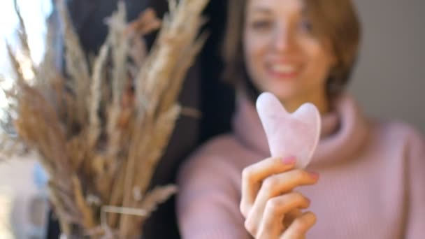 La chica muestra un masajeador de gua sha de masaje hecho de cuarzo rosa de piedra natural que se utiliza para estimular el sistema linfático y tiene un efecto anti edad, la cara femenina y las espiguillas no están en foco — Vídeo de stock