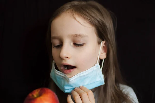 Little blonde girl with blue eyes wearing disposable mask for protection of Coronavirus Covid-19 is eating a red apple to boost her immune system during epidemic of the virus — Stock Photo, Image
