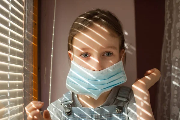 Little girl is sitting on the windowsill and trying to put protective disposable mask on her face during self-isolation at home because of Coronavirus Covid-19 on blinds background — Stock Photo, Image