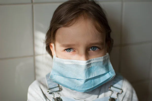 Retrato de la niña triste en una máscara protectora desechable se queda en casa debido a la pandemia de Coronavirus Covid-19 en auto-aislamiento sobre fondo de baldosas blancas en la cocina durante el día soleado —  Fotos de Stock