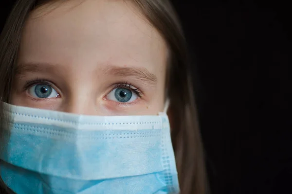 Retrato de cerca de la niña indefensa sobre fondo negro en máscara desechable azul para la protección del Coronavirus Covid-19 durante el autoaislamiento en casa — Foto de Stock