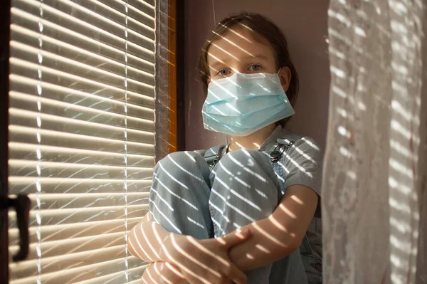 Tired bored little girl is sitting on the windowsill in protective disposable mask on her face during self-isolation at home because of Coronavirus Covid-19 on blinds background. — Stock Photo, Image
