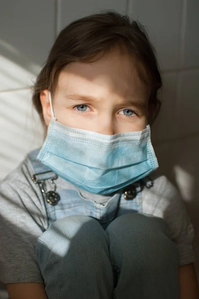 La niña triste está restringida en casa debido a la pandemia del Coronavirus Covid-19 y el autoaislamiento. Niño está sentado en la cocina sobre fondo de baldosas blancas en una máscara desechable protectora —  Fotos de Stock