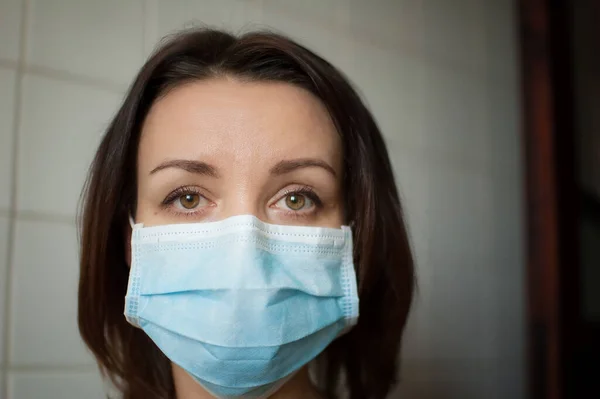 Sad adult girl is restricted at home because of the pandemia of Coronavirus Covid-19 and self-isolation. Woman is at the kitchen during sunny day on white tiles background in disposable mask — Stock Photo, Image