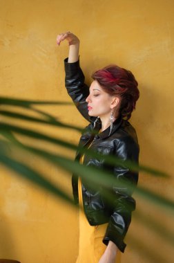 Punk rock tarzı parti kıyafetleri içinde, koyu pembe saçlı, siyah deri ceketli ve sarı arka planda uzun küpeli çekici bir kadın.