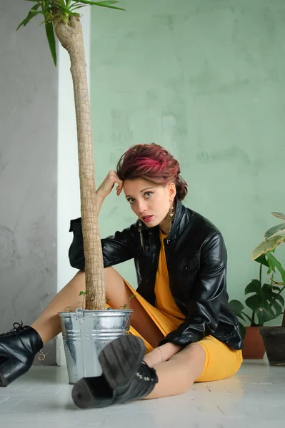 Beautiful girl in glam rock style with dark pink hair and mohawk in yellow dress and black leather jacket is sitting on the floor near a flower pot with houseplant — Stock Photo, Image