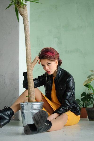 Beautiful girl in glam rock style with dark pink hair and mohawk in yellow dress and black leather jacket is sitting on the floor near a flower pot with houseplant