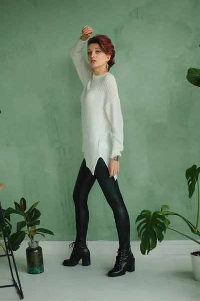 Studioporträt eines großen jungen Models in weißem Pullover und schwarzer Lederleggings, das im Studio mit grünem Hintergrund und exotischen Pflanzen posiert — Stockfoto