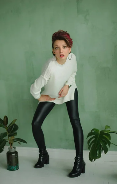 Estúdio de altura total retrato de alta jovem modelo de moda vestindo suéter branco e leggings de couro preto possuindo em estúdio com fundo verde e plantas exóticas — Fotografia de Stock