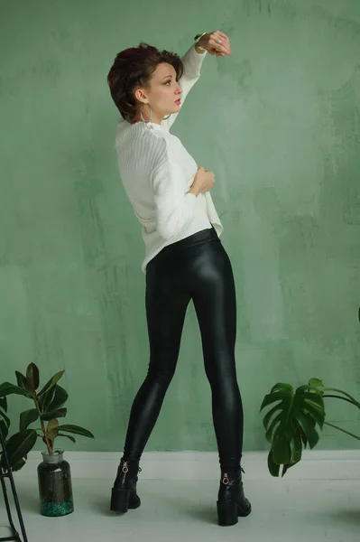 Vista posteriore di sensuale ragazza in maglione bianco e leggings in pelle nera in possesso in studio con sfondo verde e piante esotiche sul pavimento — Foto Stock