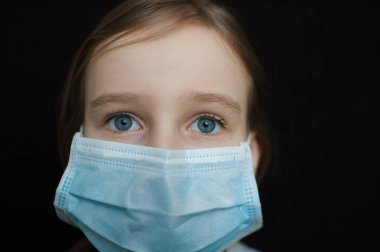 Coronavirus Covid-19 salgını. Stüdyodaki siyah arka planda virüsü korumak için mavi gözlü küçük sarışın kız tek kullanımlık maske takıyor. Kişisel koruyucu ekipman konsepti